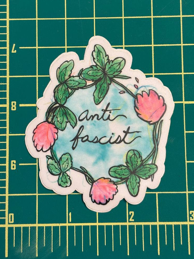 Sticker with floral design and 'anti fascist' text on a green grid background with ruler.